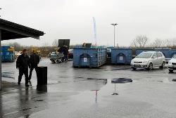 Blå containere på en regnvåd plads med to biler og to mennesker.