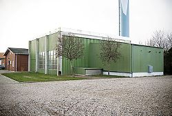 Billedet viser Hjerting Varmeværk, en stor firkantet grøn bygning med to træer og en grusplads.